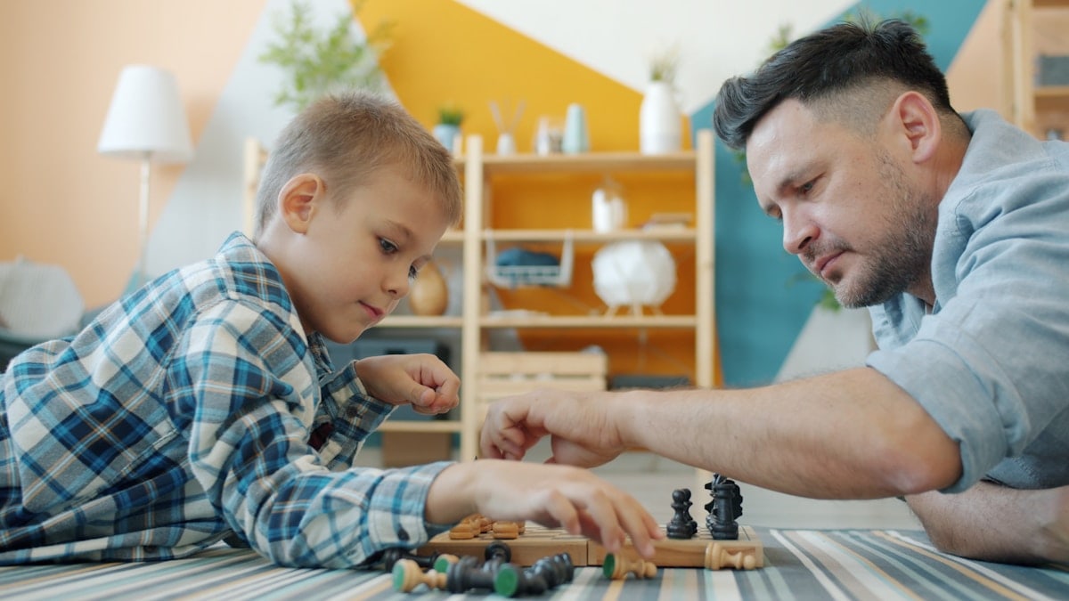 Portrait of father and son playing chess putting chessmen on board getting ready for game at home. Intellectual hobby, people and happy family concept.